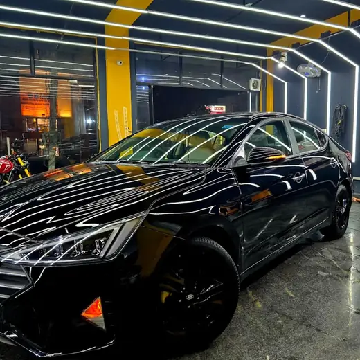 A black sedan parked in the studio bay at night, the ceiling's yellow-striped light tunnel reflected along its bonnet and roof.