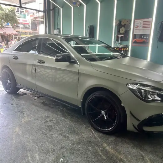 A champagne-coloured Mercedes-Benz parked side-on inside a teal-lit studio bay, with a paint-protection-film supplier's poster on the wall behind it.