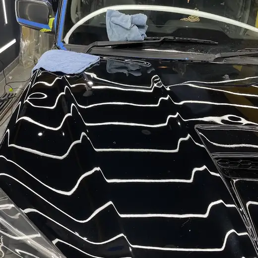 A close-up of a car's bonnet mid-installation of paint protection film, with a microfibre towel resting on the glass and blue tape wrapped around the wing mirror.